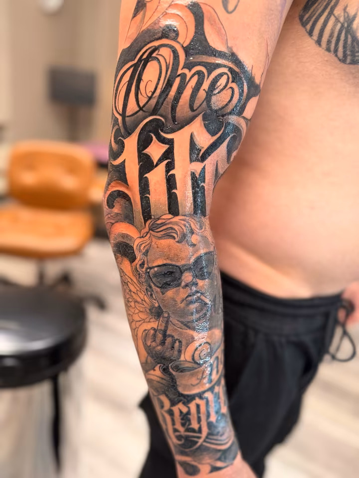 Chicano-Sleeve am ganzen Arm: Schriftzug in Script und ein Engelsmotiv in Black & Grey